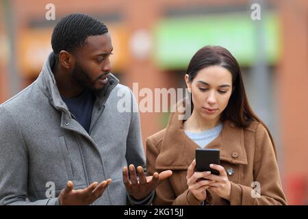 Eine Frau, die im Winter ihr Smartphone benutzt und ihren Freund auf der Straße ignoriert Stockfoto