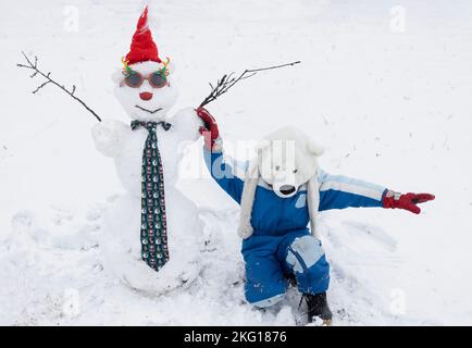 Ein nicht wiedererkennbarer kleiner Junge mit Eisbärmütze und ein Schneemann mit Weihnachtsmannmütze, Rentierbrille und Neujahrsschlips. Festliche Weihnachtszeit, ch Stockfoto