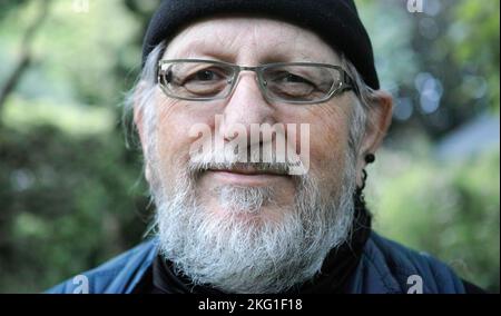 Porträt eines 68-jährigen älteren Mannes in blauer Kleidung und schwarzer Mütze. Er hat einen Bart, eine Brille und schwarze Ohren. Der Hintergrund ist unscharf Stockfoto