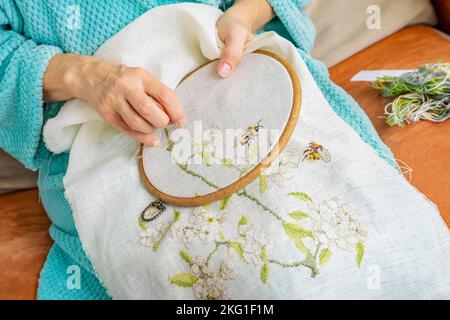 Hände einer Seniorin, die Stickerei in einem hölzernen Stickereifen auf dem Sofa macht Stockfoto