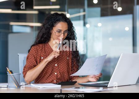 Schöne hispanische Frau im Büro, die Papierkram macht, erfolgreiche Geschäftsfrau Finanzierin, die im Büro am Tisch mit Laptop sitzt, bei der Arbeit mit Verträgen und Rechnungen, über Entscheidungen nachdenkt. Stockfoto
