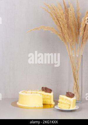 Die Untertasse ist ein Stück Kuchen aus einem gelben Cremebrot mit Schokoladenrosen und einem Strauß getrockneter Schilfblumen in einer Glasvase auf grauem Hintergrund W. Stockfoto