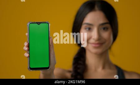 Indische ethnische Frau Dame Mädchen Dame in gelben Studio-Hintergrund zeigt Smartphone Handy-Bildschirm mit grünen leeren Chroma-Taste Touchscreen Stockfoto
