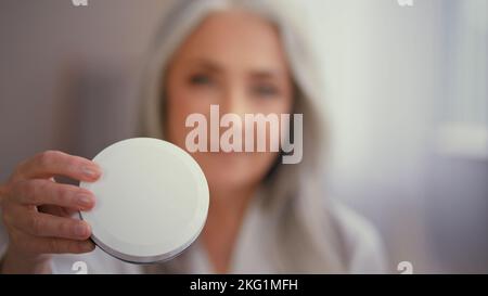 Alte kaukasische 50s ältere Frau Großmutter Dame mit langen grauen Haaren im Bademantel zeigt Glas von natürlichen organischen Sonnencreme Mineralcreme mit Stockfoto