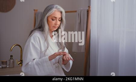 Schöne Oma 60s Dame alt Senior grauhaarige weibliche kaukasische Frau im Schlafzimmer reiben faltige Hände Anwendung natürliche Handcreme feuchtigkeitsspendend Stockfoto