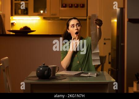 Hausbuchhaltung. Schockierte junge kaukasische Frau sitzt am Tisch und überprüft die Ausgaben auf der Quittung. Abend zu Hause. Hausfrau ist in BU beschäftigt Stockfoto