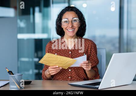 Glückliche hispanische Frau lächelnd und mit Blick auf die Kamera erhielt eine Geschäftsfrau einen Umschlag mit einem guten Nachrichtenbrief, eine Büroangestellte mit Dokumenten und einem Laptop, die im Büro bei der Arbeit arbeitete. Stockfoto