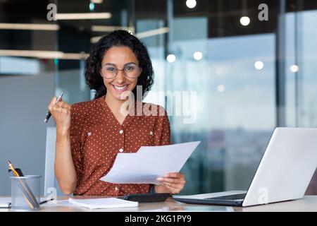 Erfolgreiche und glückliche Geschäftsfrau zufrieden mit den Erfolgen, lächelnd und mit Blick auf die Kamera, die Dokumente und Finanzberichte hält, hispanic-Frau, die im Büro am Arbeitsplatz mit einem Laptop arbeitet. Stockfoto
