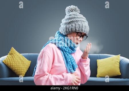 Die junge Frau in Brille, Hut und Strickschal wärmt sich und hustet. Grauer Hintergrund mit Sofa. Das Konzept von Krankheit, Grippe und Winter Stockfoto