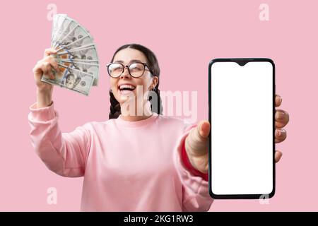 Investitionen und Handel an der Börse. Kaukasische fröhliche junge Frau mit Brille zeigt einen Geldfan und hält ein Smartphone mit weißem Scre Stockfoto