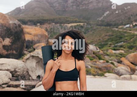 Porträt einer Frau mit lockigem Haar mit Yogamatte, die draußen an den Bergen steht Stockfoto