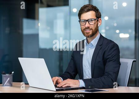 Porträt eines reifen erfolgreichen und glücklichen Geschäftsmannes, eines älteren Mannes mit lächelndem Bart und Blick auf die Kamera, eines Investors, der mit einem Laptop im Büro im Geschäftsanzug arbeitet. Stockfoto