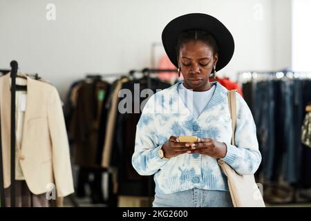 Waist-up-Porträt einer jungen schwarzen Frau mit Smartphone beim Einkaufen im Spargeschäft, Kopierraum Stockfoto