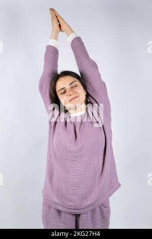 Junge Brünette Frau in Musselin Lavendel Schlafanzug Schlafanzug erstreckt sich, indem sie ihre Arme hochhebt und ihre Augen schließt Stockfoto