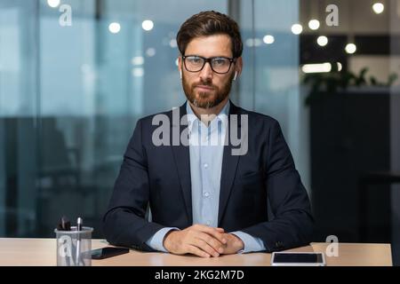 Porträt eines reifen Chefs in einem Geschäftsanzug, ein Geschäftsmann sitzt an einem Schreibtisch und schaut auf die Kamera, ein älterer Mann arbeitet im Büro. Stockfoto