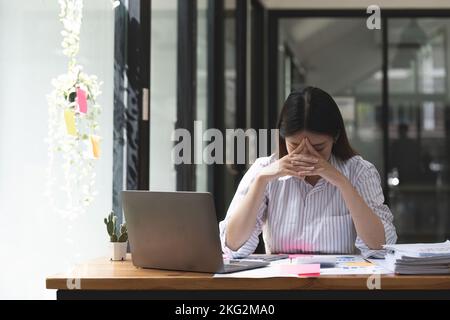 Asiatische Frauen sind gestresst, wenn sie mit einem Laptop arbeiten, müde asiatische Geschäftsfrau mit Kopfschmerzen im Büro, sich krank fühlen bei der Arbeit Stockfoto