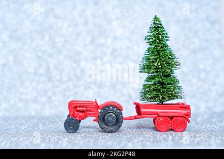 Spielzeugtraktor mit einem kleinen Weihnachtsbaum isoliert auf glitzerndem Hintergrund. Anbau von Kiefern, Fichten und Tannen für die Feiertage. Stockfoto