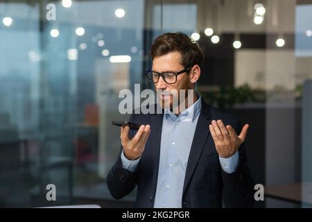 Freundlicher und lächelnder Geschäftsmann im Büro, der Audio-Nachrichten aufzeichnet, Mann, der Smartphone-App für die Kommunikation nutzt, Investor in Geschäftsanzug in der Nähe des Fensters im Gebäude. Stockfoto