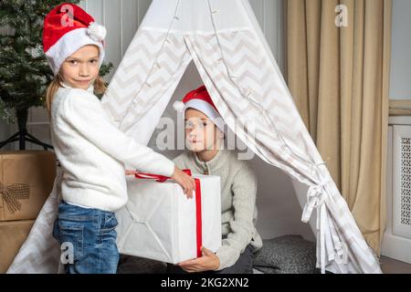 Ein Mädchen in einem Weihnachtsmütze schenkt einem Jungen zu Weihnachten und Neujahr im Kinderzimmer. Bruder und Schwester tauschen Geschenke aus. Kinder schenken Geschenke. Frohe Stockfoto