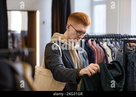 Junger rothaariger Mann, der Kleidung auf Regalen durchstöbert, während er im Spargeschäft nachhaltig einkauft Stockfoto