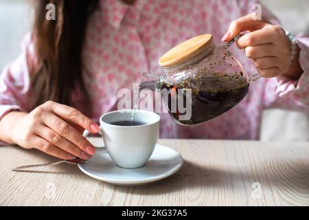 Frau weibliche Hände halten transparente Teekannen mit Kräutertee gesund wärmend und gießen heißen Tee in weiße Tasse Stockfoto