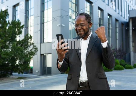 Erfolgreicher afroamerikanischer reifer Investor mit Telefon vor dem Bürogebäude, der online glückliche Nachrichten liest, ein älterer Geschäftsmann feiert den Sieg in Geschäftsanzug und Brille. Stockfoto