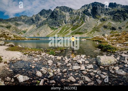 Wanderer Frau erkunden Sie die fantastische Landschaft und genießen Sie die Aussicht in der Nähe eines Bergsees in gelb in Jacke, hohe Tatra, Slowakei Stockfoto