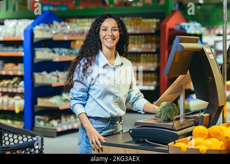 Porträt einer glücklichen Frau Shopper in einem Supermarkt wiegt eine hispanische Frau Brokkoli-Kohl auf einer Waage, lächelt und schaut auf die Kamera, in einem Geschäft zwischen Regalen mit Lebensmittelprodukten. Stockfoto