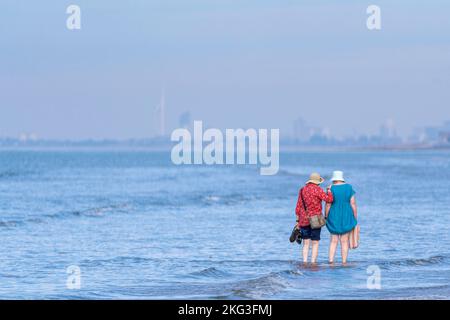 Zwei Damen paddeln im herrlichen Wasser des West Wittering Beach mit Blick auf Portsmouth's Spinnaker Tower. Stockfoto