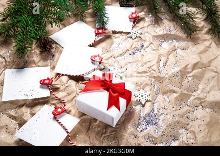 Mehrere weiße Briefpapier-Blätter zum Schreiben auf Neujahrshintergrund und eine Geschenkbox mit einer schönen roten Schleife. Frohes neues Jahr und Frohe Weihnachten. Neujahrsziele oder -Versprechen mit bunten Dekorationen auf dem Hintergrund von zerknittertem Kraftpapier mit Christbaumzweigen. Hoher Winkel. Stockfoto