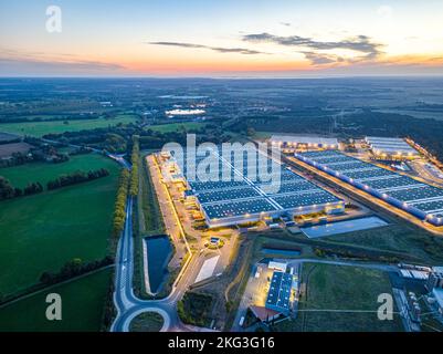 Blick aus der Vogelperspektive auf das abendliche Lagerhaus in der Nähe der Autobahn, umgeben von üppigem Grün und Lastwagen, die in der Laderampe im Val d'Oise, Frankreich, geparkt sind Stockfoto