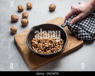 Eine Frau hält den Griff einer schwarzen Pfanne mit gerösteten Walnüssen. Betonhintergrund. Stockfoto