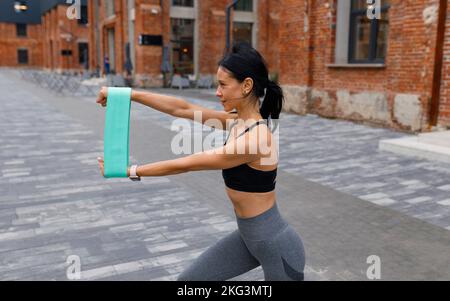 Eine attraktive sportliche Frau mit elastischen Fitnessbändern in den Händen macht Ausfallschritte auf der Straße Stockfoto