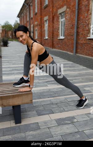 Die fröhliche brünette Frau in dunkler Sportbekleidung dehnt vor dem Laufen ihre Beinmuskeln auf einer Stadtbank Stockfoto