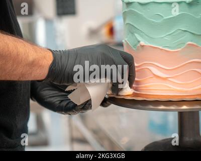 Kuchendesigner glättet einen mattierten, leeren Kuchen mit türkisfarbener, hellblauer und pinkfarbener Buttercreme mit Spachtel Stockfoto