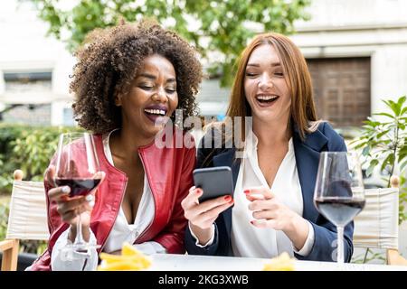 Zwei Erwachsene Frauen, die sich während der Happy Hour nach der Arbeit amüsieren, und Kollegen, die lachen, während sie sich ein Smartphone, ein soziales Netzwerk und eine mobile App anschauen Stockfoto