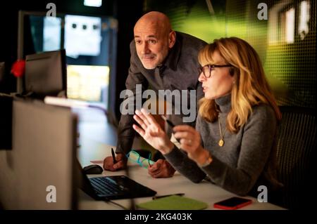 Eine Gruppe ungezwungener Geschäftsleute, die am späten Abend im Büro mit Computern am Tisch sitzen und geschäftliche Informationen analysieren. Teamarbeit. Stockfoto