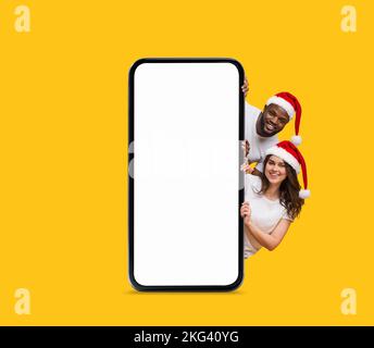 Glückliches schwarzes und europäisches junges Paar in Weihnachtsmann-Hüten, das aus einem großen Smartphone mit leerem Bildschirm guckt Stockfoto