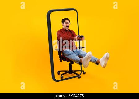 Ein glücklicher, überraschter junger europäischer Gamer mit offenem Mund in Brille sitzt auf einem Stuhl mit Joystick und spielt im Online-Spiel Stockfoto