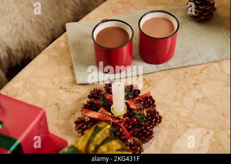 Nahaufnahme von roten Tassen mit heißem Schokoladengetränk auf grüner Serviette, neben weihnachtlich beleuchteter Kerze und Geschenken auf Marmortisch Stockfoto