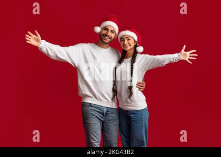 Ein Fröhliches Arabisches Paar, Das Weihnachtsmannmützen Trägt, Hände Verteilt Und Die Kamera Anlächelt Stockfoto