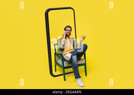 Ein lächelnder europäer der Generation Y mit Brille sitzt auf einem riesigen Smartphone-Bildschirm und telefoniert in einem Sessel Stockfoto