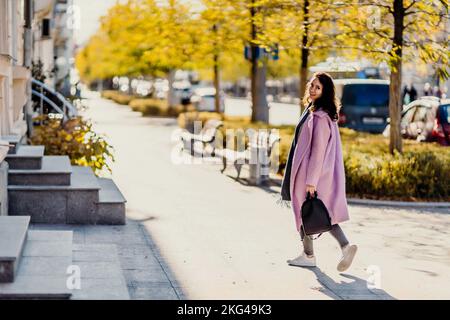 Frau Herbststadt. Eine Frau in einem rosa Kunstpelzmantel, die im Herbst an einem sonnigen Tag auf einer Straße posiert. Bäume mit gelben Blättern entlang der Straße. Stockfoto