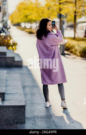 Frau Herbststadt. Eine Frau in einem rosa Kunstpelzmantel, die im Herbst an einem sonnigen Tag auf einer Straße posiert. Bäume mit gelben Blättern entlang der Straße. Stockfoto