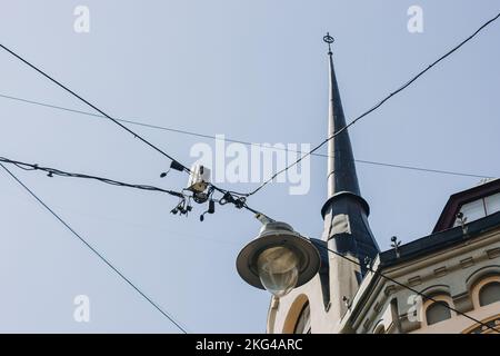 Nahaufnahme von Straßenlaternen, die in x-förmigen Drähten unter einem blauen Himmel neben einem riesigen Wohnhaus mit einem turmartigen Dach installiert sind. Stockfoto