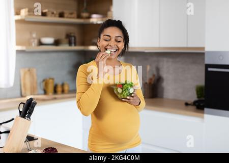 Glückliche, schwarze, schwangere Frau mit großem Bauch, die Bio-Gemüse aus Salat isst Stockfoto