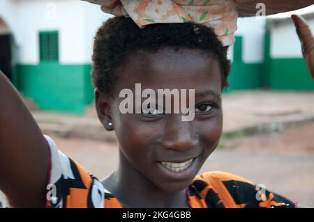 Ein junges Mädchen, das Waren zum Verkauf auf dem Kopf trägt, Natitingou, Atacora, Benin. Moschee im Hintergrund. Von früh an lernen junge Mädchen, zu tragen Stockfoto