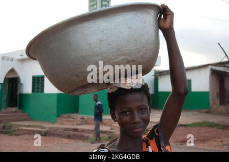 Ein junges Mädchen, das Waren zum Verkauf auf dem Kopf trägt, Natitingou, Atacora, Benin. Moschee im Hintergrund. Von früh an lernen junge Mädchen, zu tragen Stockfoto