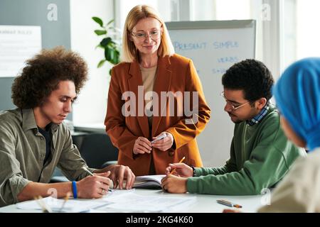 Selbstbewusster Lehrer, der vor einer Gruppe junger interkultureller Studenten steht, die sich in ihren Copybooks während einer Lektion oder eines Seminars Notizen machen Stockfoto