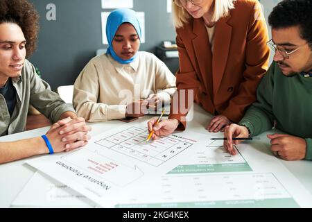 Reifer blonder Lehrer, der sich über Dokument mit Pronomen beugt, während er der Gruppe interkultureller Schüler neues Thema erklärt Stockfoto